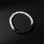 A beaded stretch bracelet arranged in a circle on a dark background, featuring translucent white round beads on one side and matte black porous round beads on the other, with a small metallic silver bead near the top right.