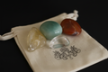 Four polished tumbled crystals in yellow, light green, reddish-brown, and clear, displayed on a cream drawstring pouch with a black background.
