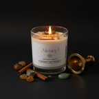 A lit white candle in a clear glass jar with a flame, styled on a dark surface with crystals, cinnamon sticks, and a brass bell. The label reads 'Money Intention Candle'.