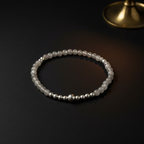 A beaded stretch bracelet featuring white translucent beads and small metallic silver beads, displayed on a dark surface with a blurred gold object in the background.