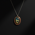 A copper oval pendant necklace featuring a natural labradorite stone with a blue and green iridescent sheen, suspended from a silver-toned chain against a dark background.