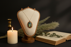 An oval pendant necklace displayed on a cream fabric jewelry bust with a wooden frame, styled with a lit white candle, an open book, dried grass, and green leaves against a dark background.