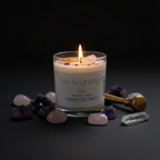 A lit candle in a clear glass jar with a cream label reading 'New Beginnings Intention Candles,' styled on a dark surface with pink and purple crystals and a brass tool around it.