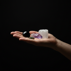 A human hand holding several small crystals and stones against a solid black background, including a black sphere, a white cuboid crystal, a purple amethyst cluster, and a small green stone.