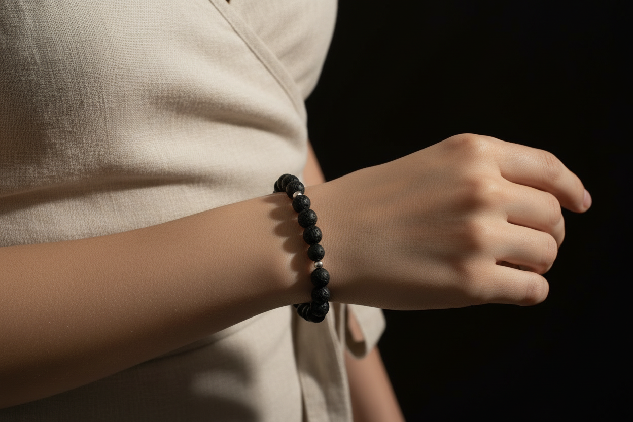 A close-up of a person's wrist wearing a dark beaded bracelet, with a light beige linen top and a solid black background.