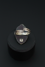 A sterling silver ring with a unique organic-shaped bezel setting, displayed on a black cylindrical stand against a dark background. The ring features a split band design and a small engraved 'JM' mark on the bottom of the setting.