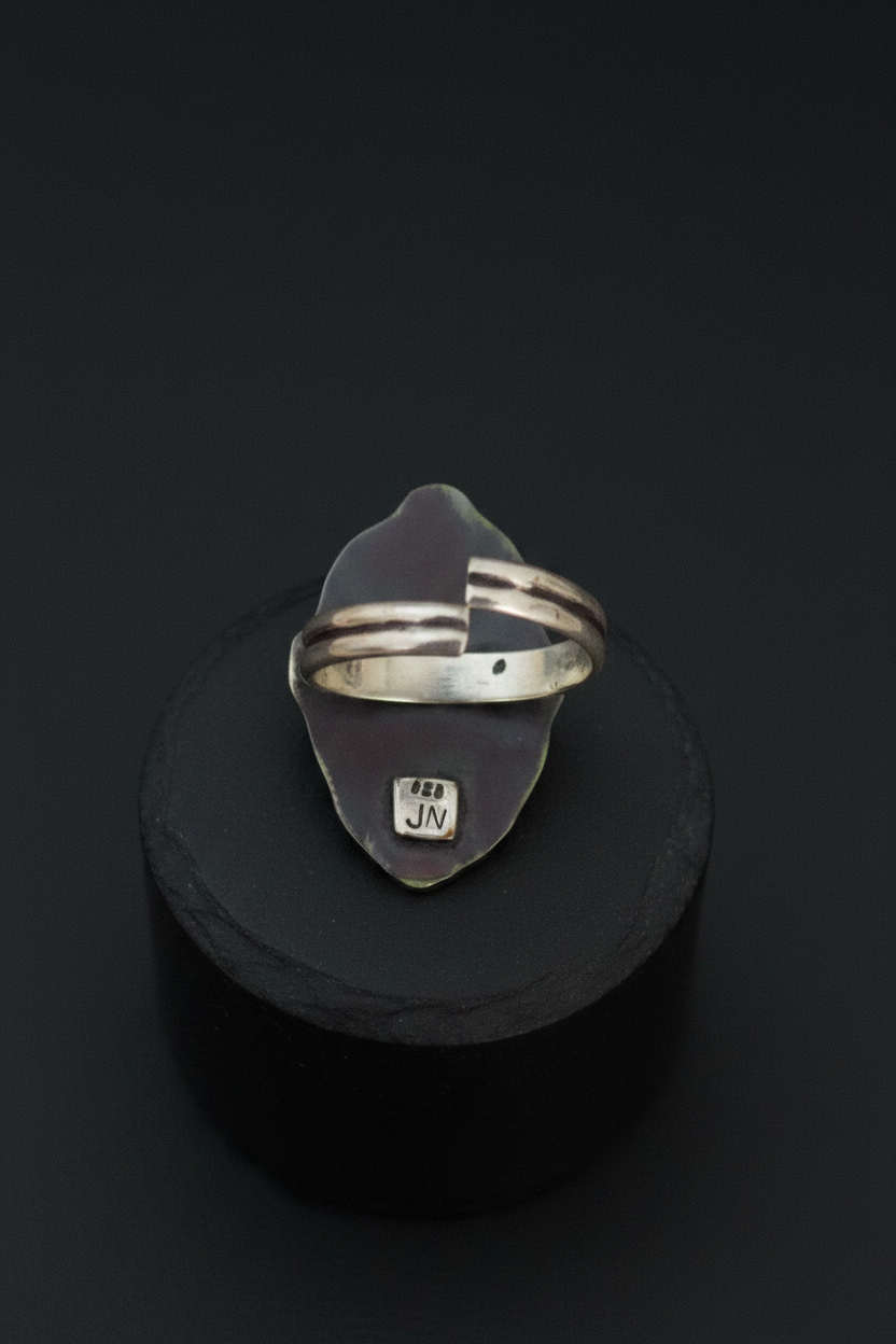 A sterling silver ring with a unique organic-shaped bezel setting, displayed on a black cylindrical stand against a dark background. The ring features a split band design and a small engraved 'JM' mark on the bottom of the setting.