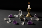 A silver ring with a large oval amethyst cluster setting, displayed on a dark surface surrounded by raw purple crystals, clear quartz points, and a lit brass candle holder against a black background.