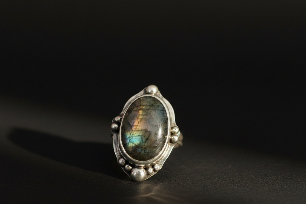 A sterling silver ring featuring an oval cabochon labradorite gemstone with a shimmering green, blue, and brown play-of-color, set against a dark, reflective background.