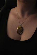 A close-up of a person wearing a silver-toned chain necklace with an oval pendant featuring a yellow-brown stone with dark inclusions, set against a dark black top.