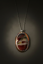 An oval pendant necklace featuring a polished red, white, and black agate stone set in a silver-toned bezel, suspended from a thin silver chain against a dark gray background.