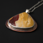 A close-up of a triangular pendant necklace featuring a marbled stone with yellow, cream, and reddish-brown sections, set in a silver-toned metal bezel with a copper-toned outer rim, hanging from a silver chain against a dark background.