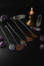 Four silver-toned necklaces with oval iridescent pendants engraved with zodiac symbols are arranged on a black surface alongside crystals, a brass candlestick with a lit candle, and purple amethyst stones.