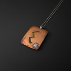 A rectangular copper pendant necklace with a black engraved zigzag design and a small silver square charm, suspended on a dark chain against a black background.