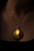 A close-up of a person's neck and chest wearing a teardrop-shaped pendant necklace. The pendant has a warm yellow and orange marbled interior with a dark outer edge and a silver-toned metal setting, suspended from a thin chain.