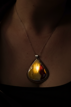 A close-up of a person's neck and chest wearing a teardrop-shaped pendant necklace. The pendant has a warm yellow and orange marbled interior with a dark outer edge and a silver-toned metal setting, suspended from a thin chain.
