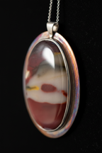 An oval pendant necklace featuring a natural marbled agate stone with shades of red, white, and gray, set in a silver-toned bezel with an iridescent rainbow edge, hanging from a silver chain against a black background.