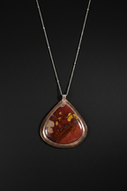 A teardrop-shaped pendant necklace featuring a polished red and orange agate stone with yellow inclusions, set in a rose gold bezel on a delicate silver chain, presented against a dark background.