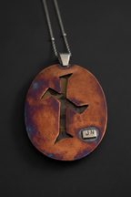 An oval pendant necklace with a rustic copper and purple-blue patina finish, featuring a cutout cross design and a small silver 'JN' nameplate, shown on a dark background.