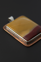 A square pendant with a polished amber-yellow and dark red banded stone face, set in a copper-toned metal frame with a silver bail, shown on a dark background.