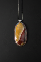 An oval pendant necklace featuring a marbled agate stone with yellow, cream, and reddish-brown patterns, set in a silver-toned bezel on a dark background.