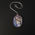 A silver necklace with an oval pendant featuring a stylized brown letter 'H' on a blue, purple, and white marbled background, displayed on a black surface.