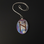 A silver necklace with an oval pendant featuring a stylized brown letter 'H' on a blue, purple, and white marbled background, displayed on a black surface.