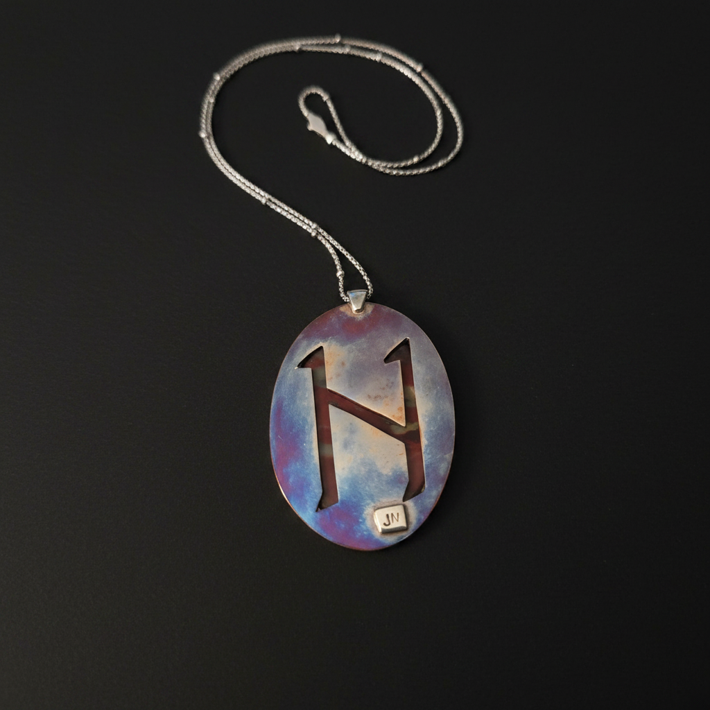 A silver necklace with an oval pendant featuring a stylized brown letter 'H' on a blue, purple, and white marbled background, displayed on a black surface.