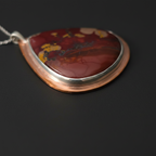 A teardrop-shaped pendant necklace featuring a polished red jasper stone with yellow and black matrix patterns, set in a copper-toned bezel with a silver-colored inner border and a silver chain.