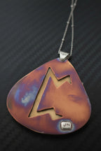 A teardrop-shaped pendant necklace with a cutout mountain symbol, featuring a copper and purple-blue oxidized finish on a silver chain, set against a dark carbon fiber background.