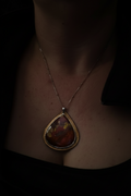 A close-up of a person wearing a gold pendant necklace with a teardrop-shaped stone featuring red, orange, and yellow marbled colors, set against a very dark background.