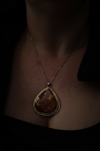 A close-up of a person wearing a gold pendant necklace with a teardrop-shaped stone featuring red, orange, and yellow marbled colors, set against a very dark background.