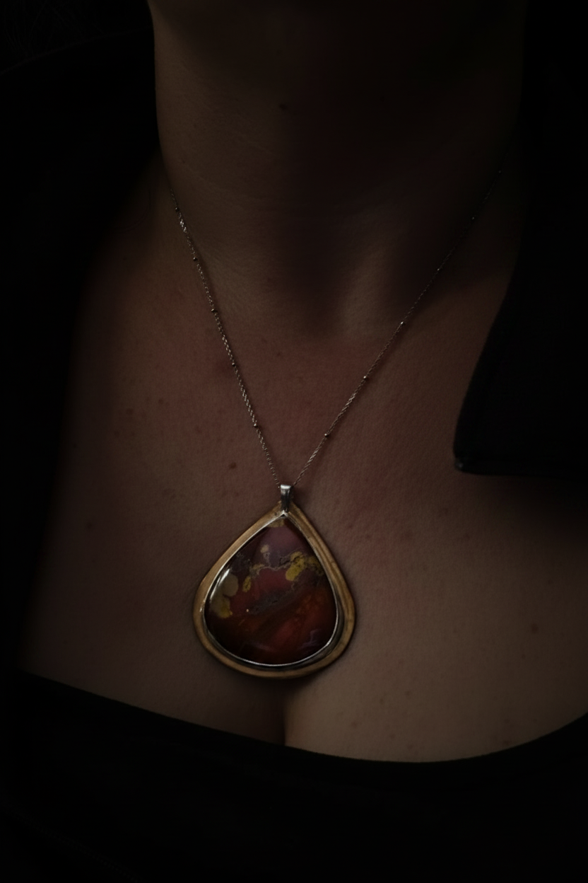 A close-up of a person wearing a gold pendant necklace with a teardrop-shaped stone featuring red, orange, and yellow marbled colors, set against a very dark background.
