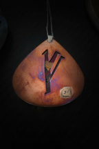 A teardrop-shaped pendant with a warm copper and orange gradient finish, featuring a large black letter 'Y' cutout in the center and a small square metallic 'JN' emblem near the bottom right. The pendant hangs from a dark cord against a solid black background.