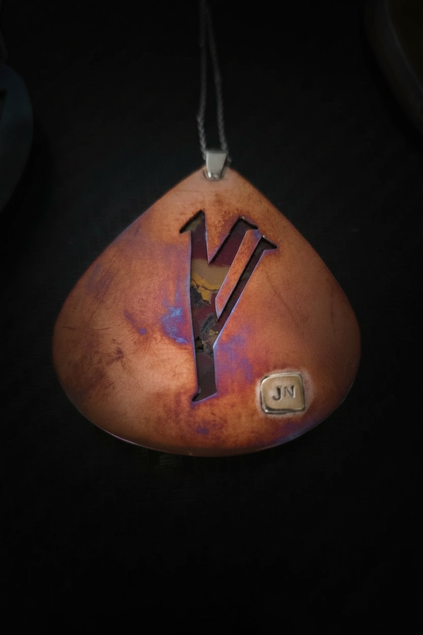 A teardrop-shaped pendant with a warm copper and orange gradient finish, featuring a large black letter 'Y' cutout in the center and a small square metallic 'JN' emblem near the bottom right. The pendant hangs from a dark cord against a solid black background.