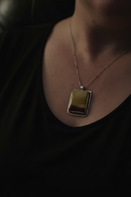 A close-up lifestyle shot of a person wearing a silver-toned rectangular pendant necklace with a yellow and dark brown gradient stone, set against a dark black top.