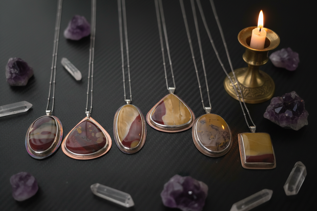 Six silver and rose gold necklaces with unique agate stone pendants are arranged on a dark textured surface alongside raw amethyst crystals and a lit brass candle holder.