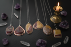 Six silver and rose gold necklaces with unique agate stone pendants are arranged on a dark textured surface alongside raw amethyst crystals and a lit brass candle holder.