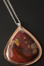 A teardrop-shaped pendant necklace featuring a polished copper-toned bezel holding a red and brown marbled stone with yellow inclusions, suspended from a silver chain against a dark background.