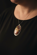 A close-up of a person wearing a delicate gold chain necklace with an oval pendant featuring a marbled brown, cream, and gold stone, set against a black top and dark background.