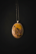 An oval pendant necklace featuring a yellow and brown marbled stone with dark inclusions, set in a silver-toned metal frame and suspended from a delicate silver chain against a solid black background.