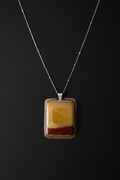 A rectangular pendant necklace featuring a layered agate or stone design with yellow, brown, and dark red bands, set in a silver-toned frame on a delicate silver chain against a dark background.