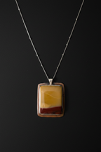 A rectangular pendant necklace featuring a layered agate or stone design with yellow, brown, and dark red bands, set in a silver-toned frame on a delicate silver chain against a dark background.