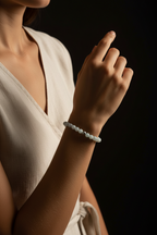 A close-up of a woman wearing a light gray beaded bracelet on her wrist, dressed in a cream-colored sleeveless top against a dark background.