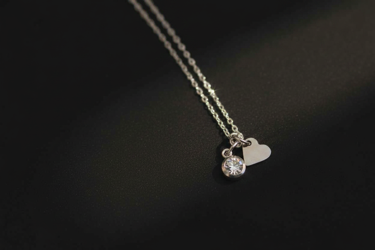 A silver chain necklace with a small round clear gemstone pendant and a small heart-shaped charm, displayed on a dark textured background.