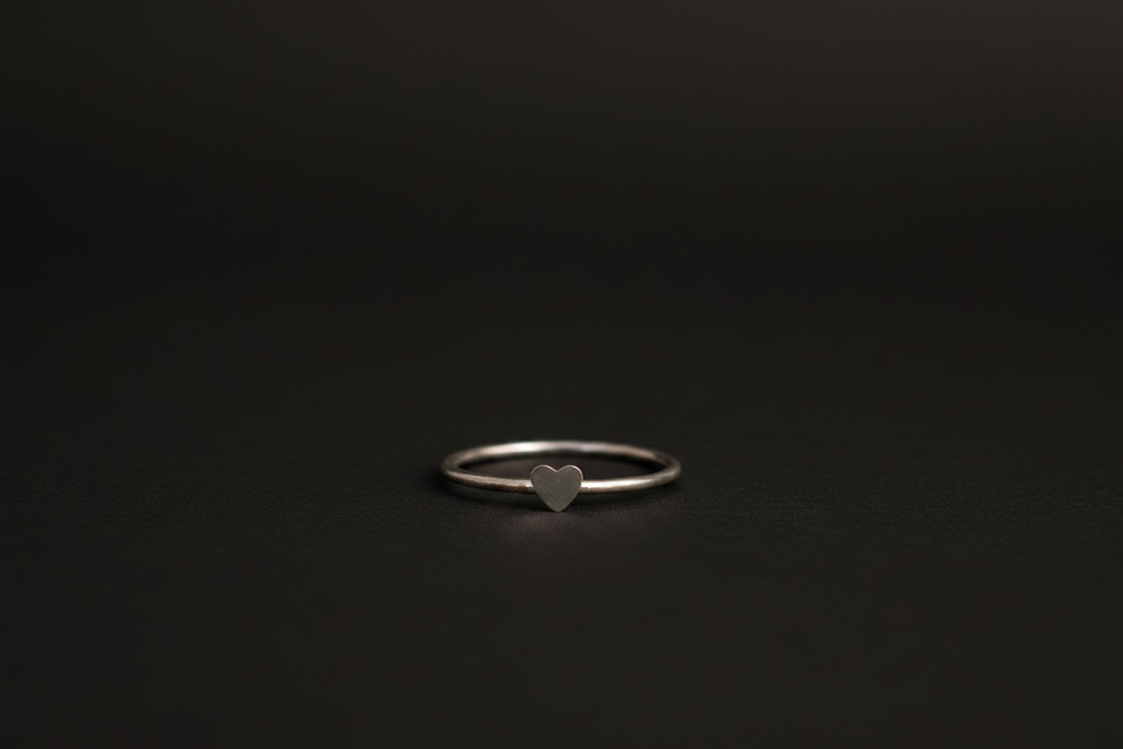 A minimalist silver band ring with a small, polished heart-shaped charm in the center, resting on a dark, textured surface.