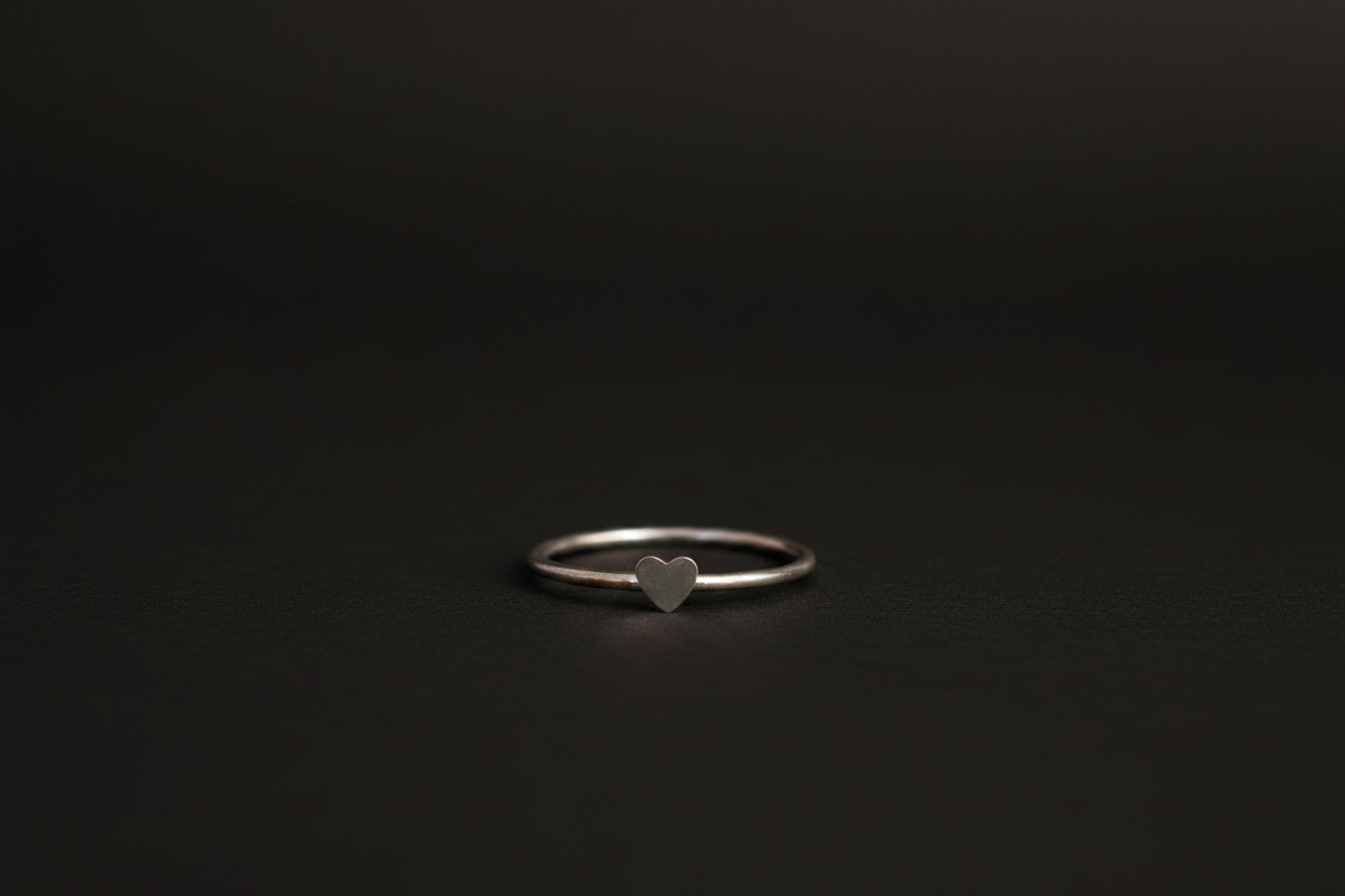 A minimalist silver band ring with a small, polished heart-shaped charm in the center, resting on a dark, textured surface.