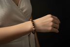 A close-up of a person's wrist wearing a beaded bracelet made of brown and gold tiger's eye beads, set against a dark background.