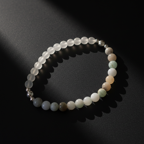 A beaded stretch bracelet made of translucent white and pale green gemstone-like beads with a few metallic silver spacer beads, photographed on a dark surface with dramatic shadows.