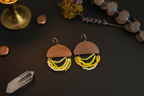 A pair of bohemian-style wooden and bead dangle earrings displayed on a dark surface with decorative props including a brass candle holder, dried lavender, and a crystal.